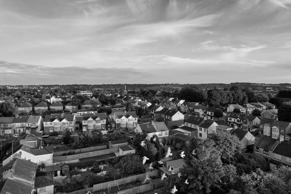 A black and white aerial photograph of a residential suburban area showing a densely packed arrangement of individual houses, many with pitched roofs and gardens, separated by fences and tree-lined st