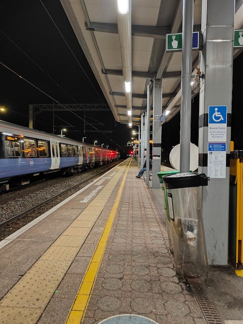 Nighttime scene at Edgware Station platform featuring a partially loaded removal van positioned adjacent to the station's shelter, with a row of cardboard boxes, wrapped furniture, and packing materials inside the vehicle indicating a home relocation process. The platform is equipped with tactile paving closest to the edge, a black and silver trash bin, and metal support columns holding a blue emergency assistance sign. A train with blue and white carriages is visible on the left, while the station's covered waiting area extends toward the background. The station lighting illuminates the platform, aiding in a safe loading and packing process conducted by Removal Van Edgware, supporting efficient furniture transport and moving logistics for residents preparing for their move near the HA8 postcode area.