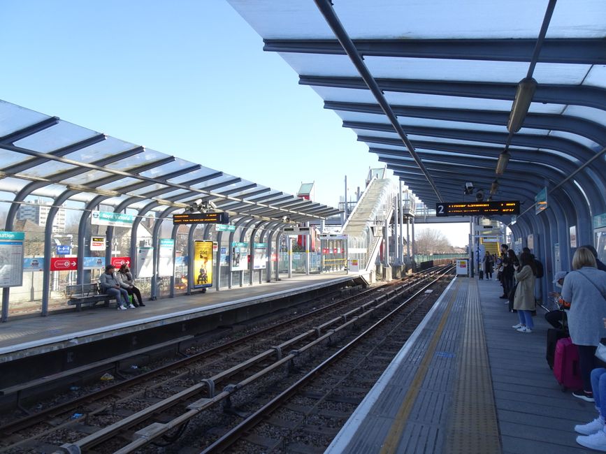 Nighttime scene at Edgware Station platform featuring a partially loaded removal van positioned adjacent to the station's shelter, with a row of cardboard boxes, wrapped furniture, and packing materials inside the vehicle indicating a home relocation process. The platform is equipped with tactile paving closest to the edge, a black and silver trash bin, and metal support columns holding a blue emergency assistance sign. A train with blue and white carriages is visible on the left, while the station's covered waiting area extends toward the background. The station lighting illuminates the platform, aiding in a safe loading and packing process conducted by Removal Van Edgware, supporting efficient furniture transport and moving logistics for residents preparing for their move near the HA8 postcode area.